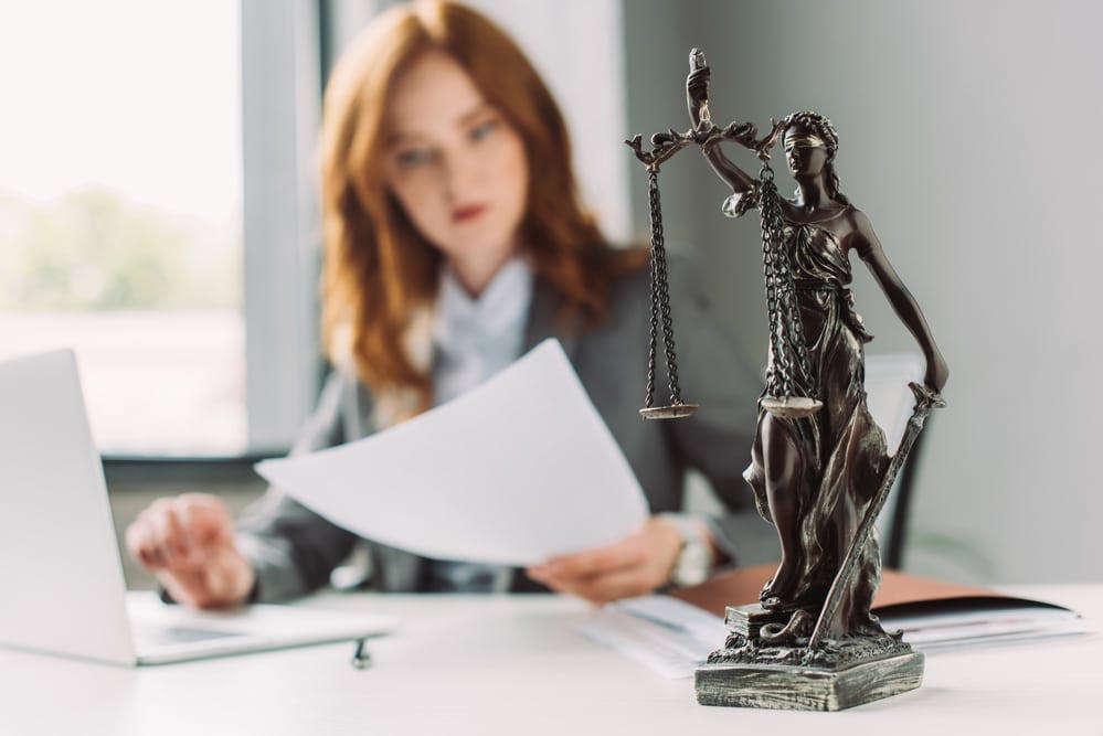 The scales of justice statute on a desk with a T-shaped lawyer in the background reviewing paperwork.